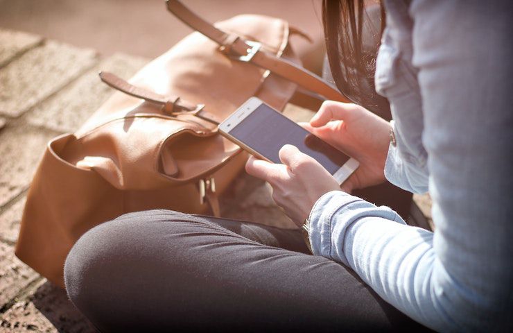 Donna che sta usando un telefono cellulare, mentre è seduta per terra davanti a una borsa