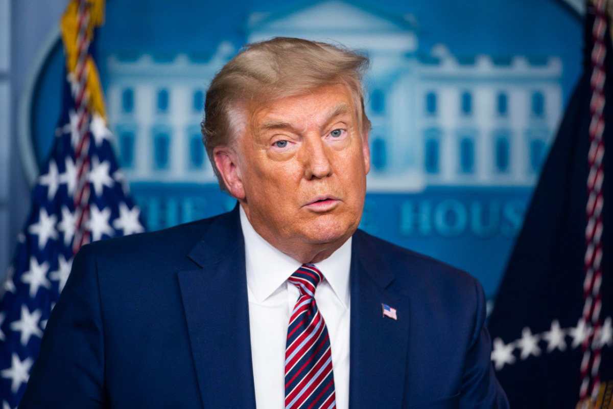 Donald Trump con giacca blu che parla con dietro una grafica della Casa Bianca